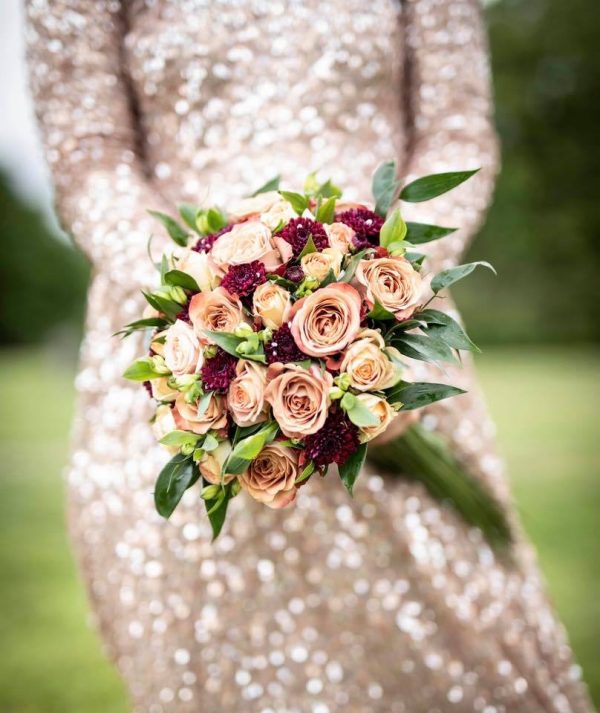 En rund brudebuket af roser og scabiosa i creme, kaffe og bourdeaux farver. Brudebuketten holdes i hånden af bruden i en glitter-kjole.