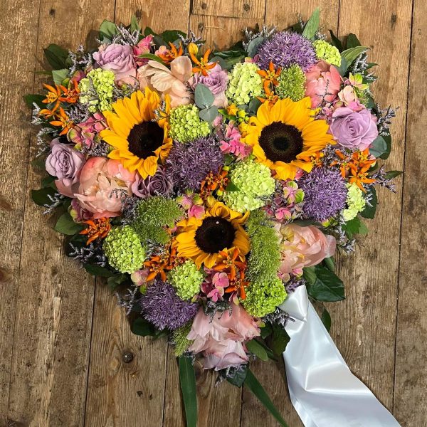 Begravelseshjerte i forskellige farver blomster og hvor der er påsat hvidt begravelsesbånd.