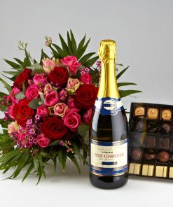 En lav tæt buket bundet af roser i blandede rodlige nuancer. En flaske bobler og en stor æske chokolade ligger ved siden af buketten.