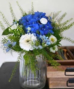 En blå og hvid buket i glasvase med en blå studenterhue og skuffer ved siden af