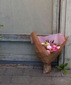 Buket af lyserøde bonderoser, pakket ind og stående udenfor ved træbaggrund.