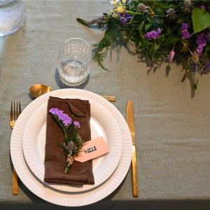 Bordpynt og tallerkenpynt i lilla farver på bord opdækket til middag med tallerken og glas, på grøn dug