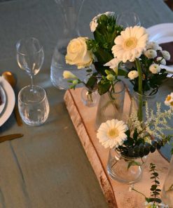 Små vaser med forskellige blomster i hvide farver placeret på en stamme af birketræ, på dækket middagsbord