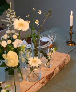 Små vaser med forskellige blomster i hvide farver placeret på en stamme af birketræ, på dækket middagsbord