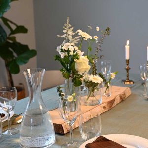 Små vaser med forskellige blomster i hvide farver placeret på en stamme af birketræ, på dækket middagsbord