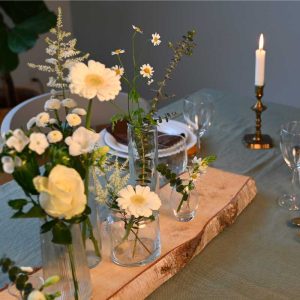 Små vaser med forskellige blomster i hvide farver placeret på en stamme af birketræ, på dækket middagsbord