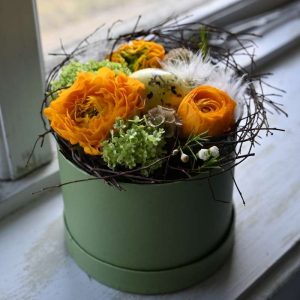 Gul påske blomsteræske med fjer og plettet påskeæg i midten.