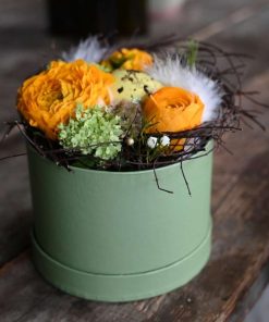 Blomsteræske med orange blomster og små grene pyntet rundt om. Et lille plettet påskeæg i midten og små hvide fjer som pynt