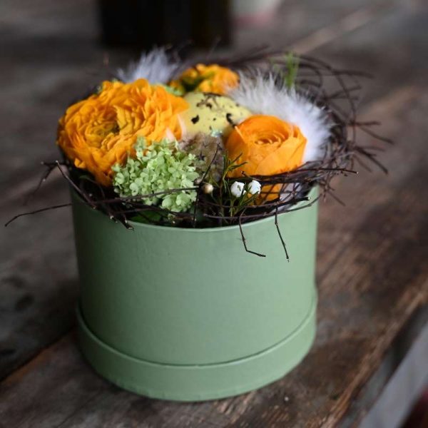 Blomsteræske med orange blomster og små grene pyntet rundt om. Et lille plettet påskeæg i midten og små hvide fjer som pynt