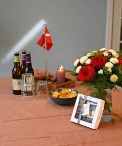 Buket i røde og hvide farver står på et bord med rosa dug, hvor der er dækket op til gæster med flag, lys, øl, chips mv.