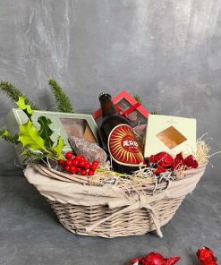 En flettet kurv med øl, slik og chokolade pyntet med julegrene og kogle og løse chokoladekugler i forgrunden.