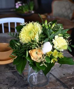 Påskebuket i sarte gule farver med hvidt påskeæg som pynt, på et rustikt bord i glasvase