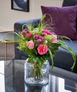 En buket i lyserøde farver på et glasbord i vase med en sofa i baggrunden