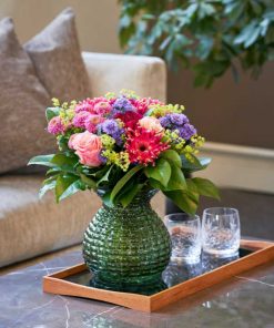 Buket i lyserøde, lilla og pinke blomster med en sofa i baggrunden og glas på et fad på bordet