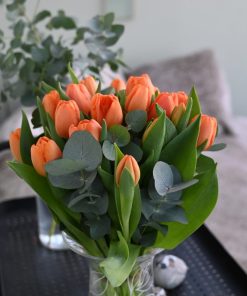 Buket orange tulipaner i en gennemsigtig glasvase, der står på et sort rullebord i hjemlige omgivelser.