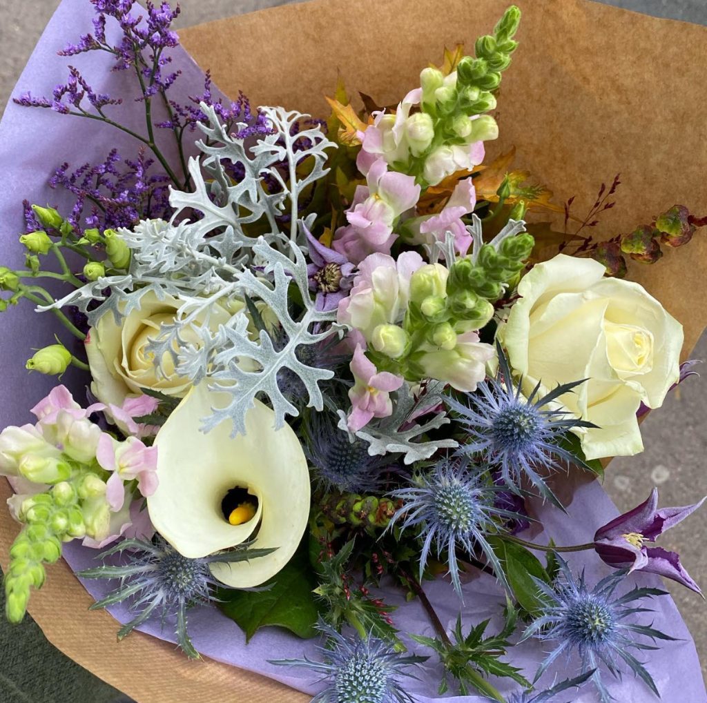 En buket blandede blomster i blå, lilla, hvid og lyserød. Buketten er pakket i brunt papir.