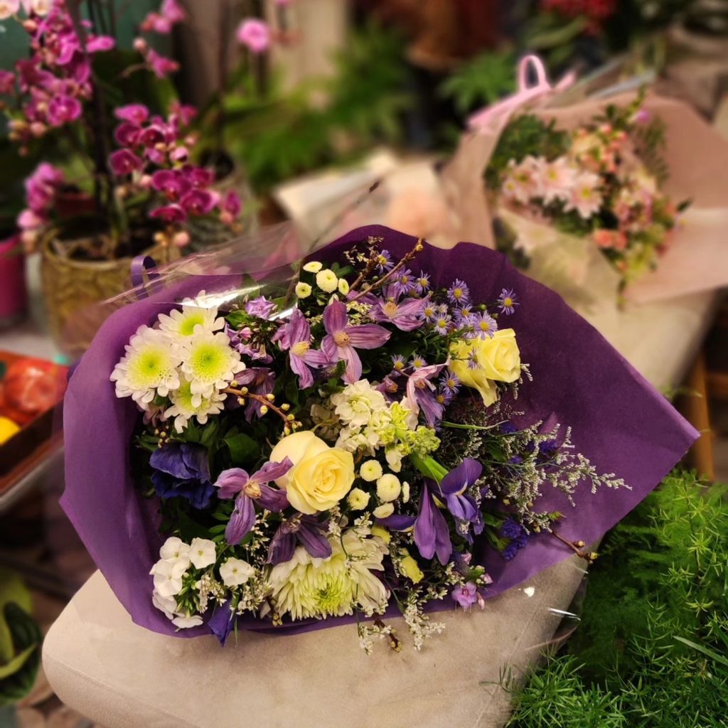 Indpakket buket i lilla og hvide. Buketten ligger på en bænk, der står i en blomsterbutik.