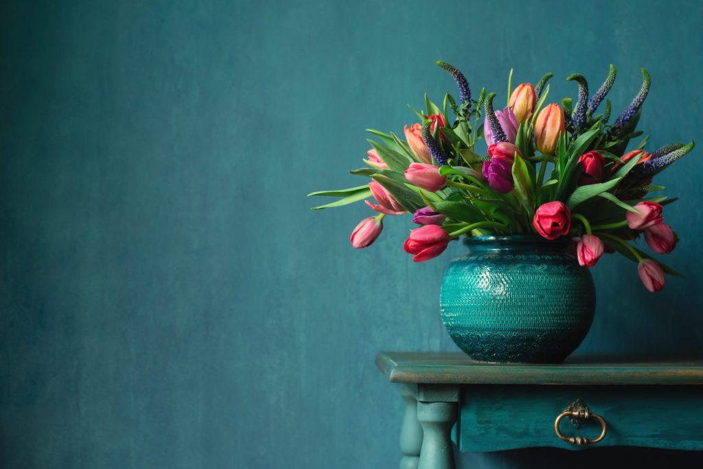 Blandede tulipaner bundet sammen med lilla veronica som står i en turkisblå, rund vase, som er placeret på et lille bord.